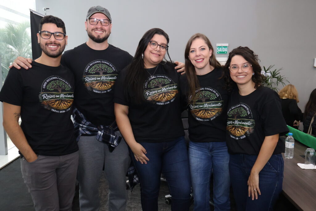Fotografia no auditório da OAB, com dois homens, brancos, e três mulheres, brancas, lado a lado posando para foto. Eles vestem camiseta preta com o logo da chapa Raízes em Movimento da seccional Caxias do Sul.
