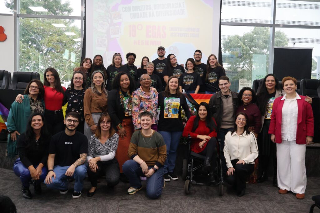 Fotografia com a nova gestão do CRESSRS, representantes da sede em Porto Alegre e das seccionais em Caxias do Sul e Pelotas. Uma pessoa não binária, 21 mulheres, sendo 1 pessoas em situação de cadeira de rodas e seis mulheres negras. Quatro homens, brancos. O grupo está no auditório da OAB, com banner do 19º EGAS atrás deles.