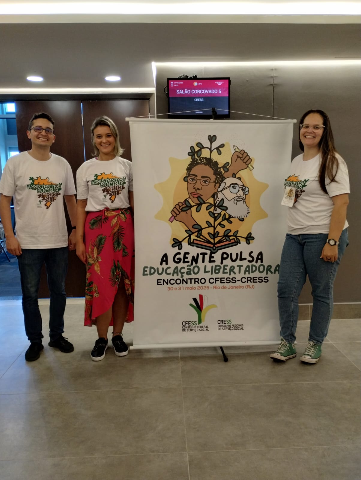 fotografia com a presença de um homem, branco, e duas mulheres brancas, que vestem a camiseta escrito assistentes sociais do Brasil e posam ao lado de um banner escrito a gente pulsa educação libertadora - encontro cfess-cress.