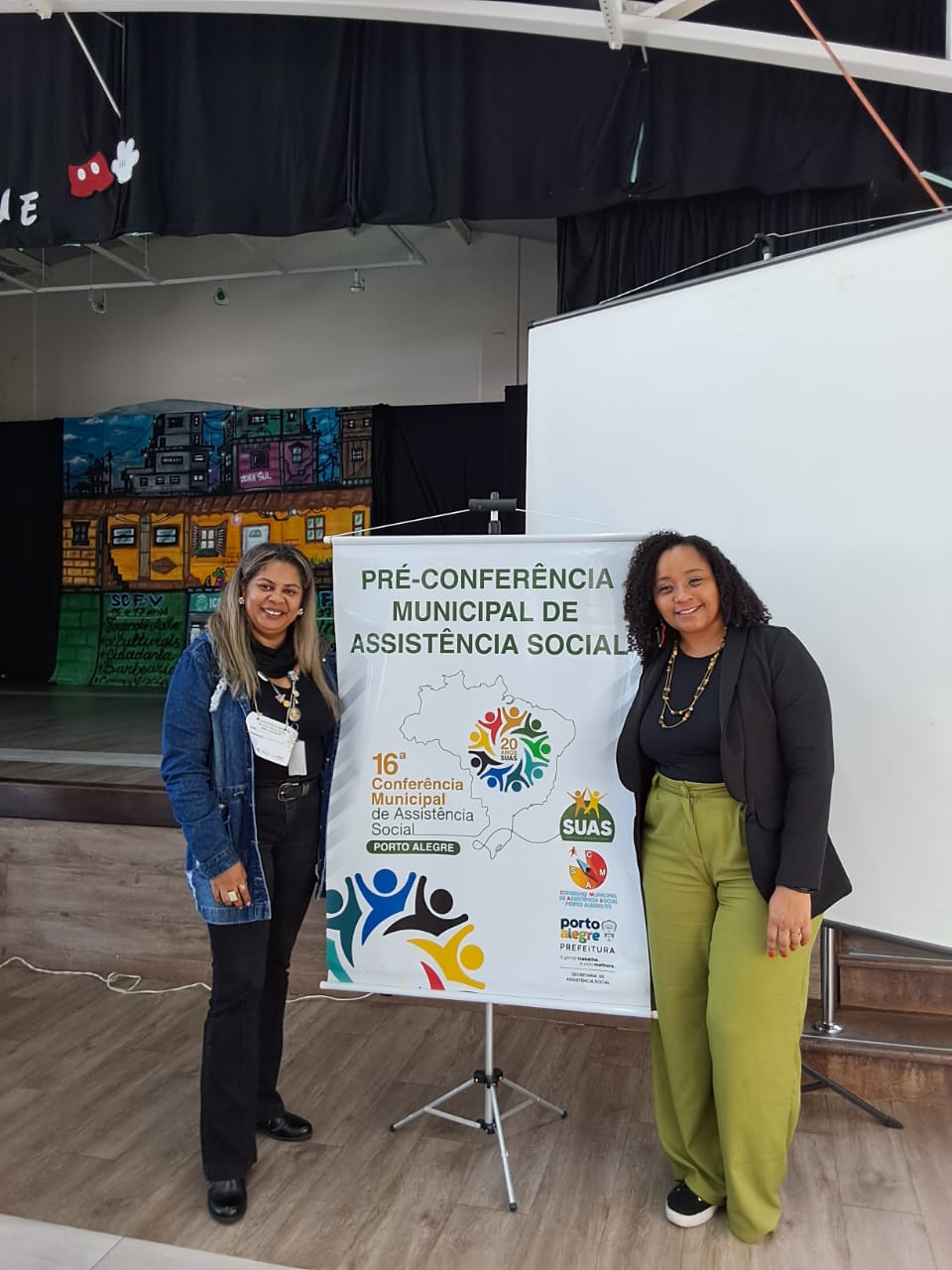 fotografia com a representante do CRESSRS, mulher, preta, e outra mulher, preta, posando ao lado do banner da pré-conferência.