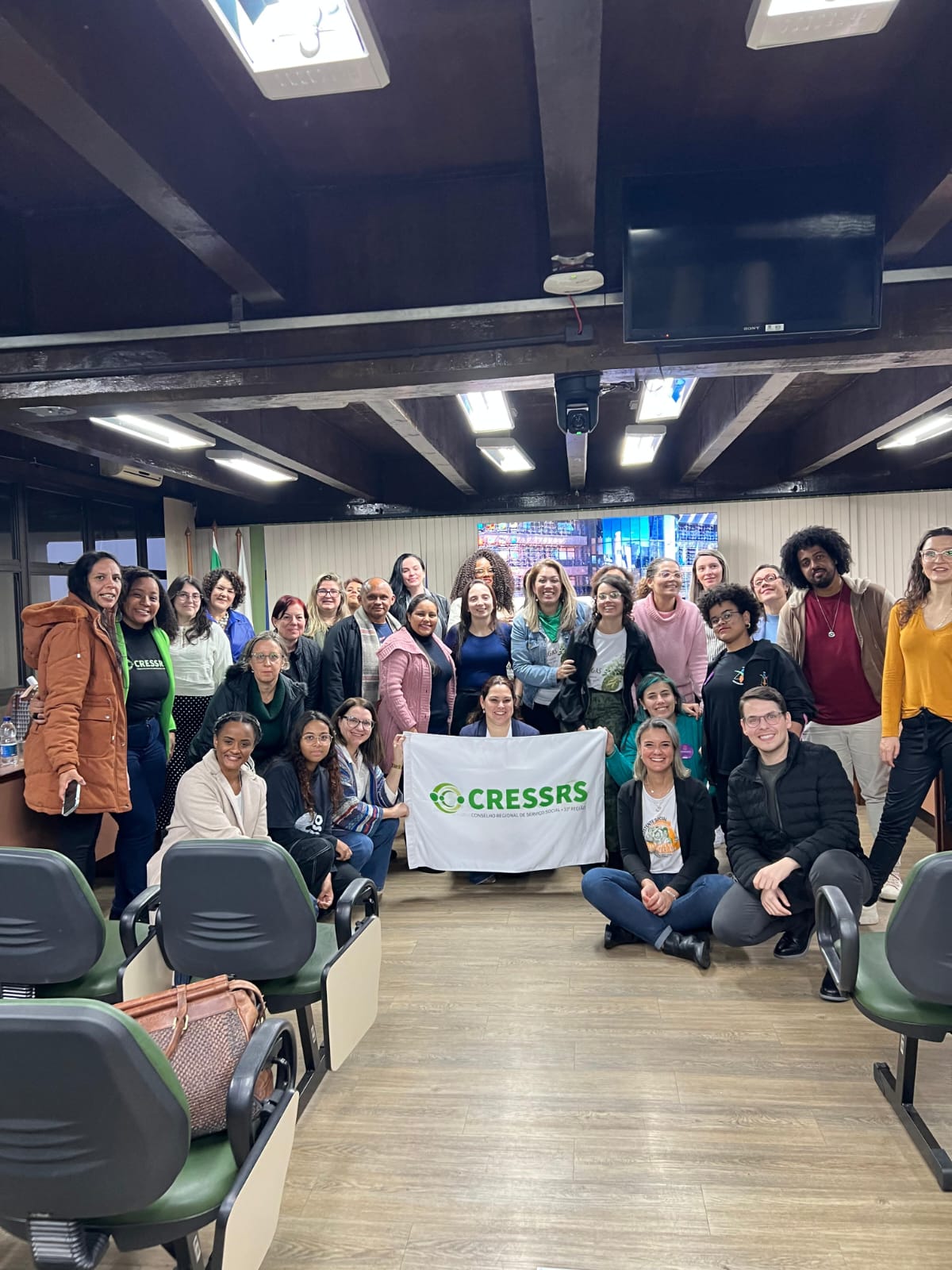 Fotografia com 27 pessoas no auditório da Câmara de Vereadores de Caxias do Sul. Ao centro da foto, uma mulher segura uma bandeira do CRESSRS. Abaixo da foto está escrito: Seccional Caxias do Sul do CRESSRS celebra 40 anos de compromisso com o Serviço Social.