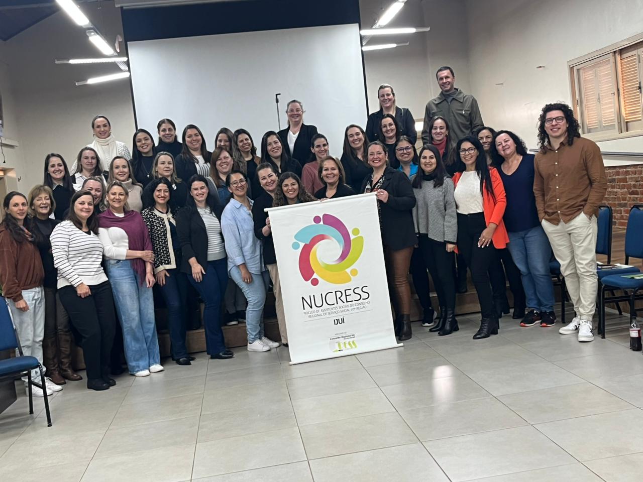 Fotografia com 37 pessoas, entre participantes do mnicurso, palestrante e representantes do Cress. A maioria das pessoas são mulheres. No centro da foto, elas seguram um banner no NUCRESS Ijuí.