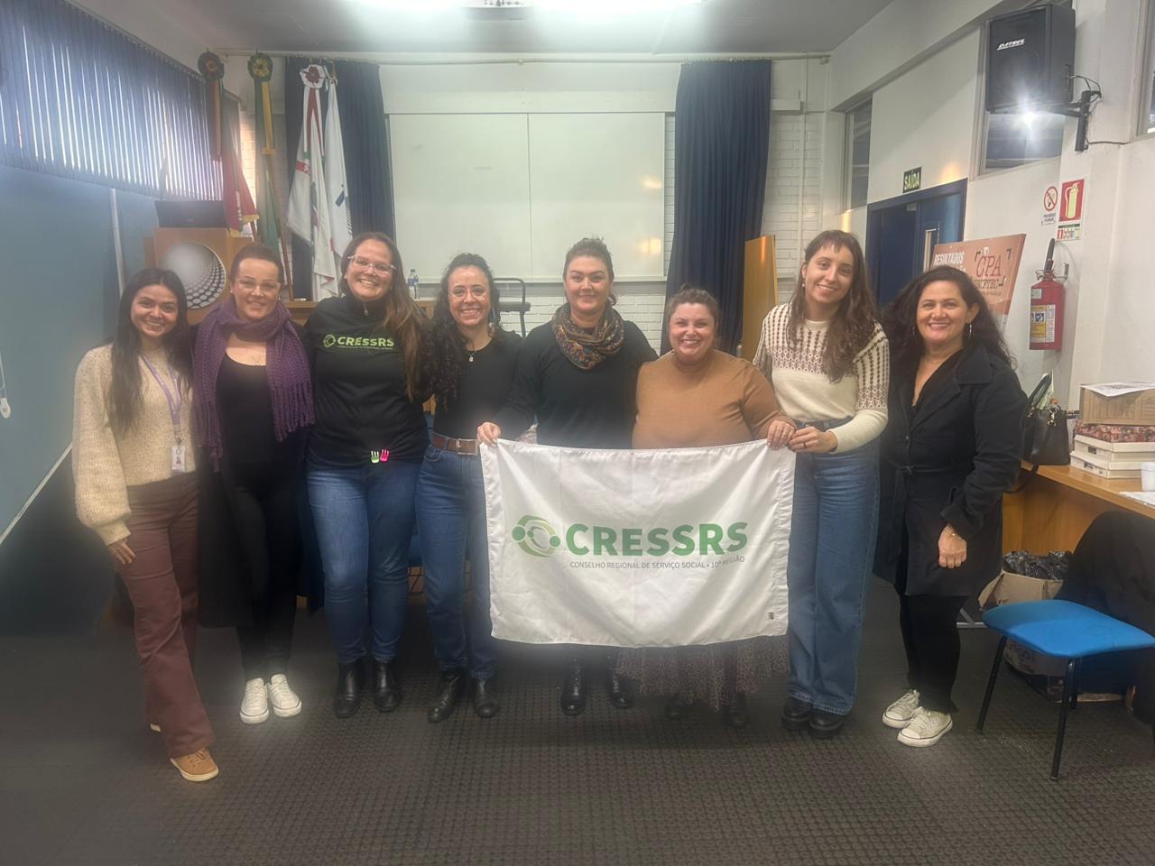Oito mulheres, brancas, seguram uma bandeira grande do CRESSRS.