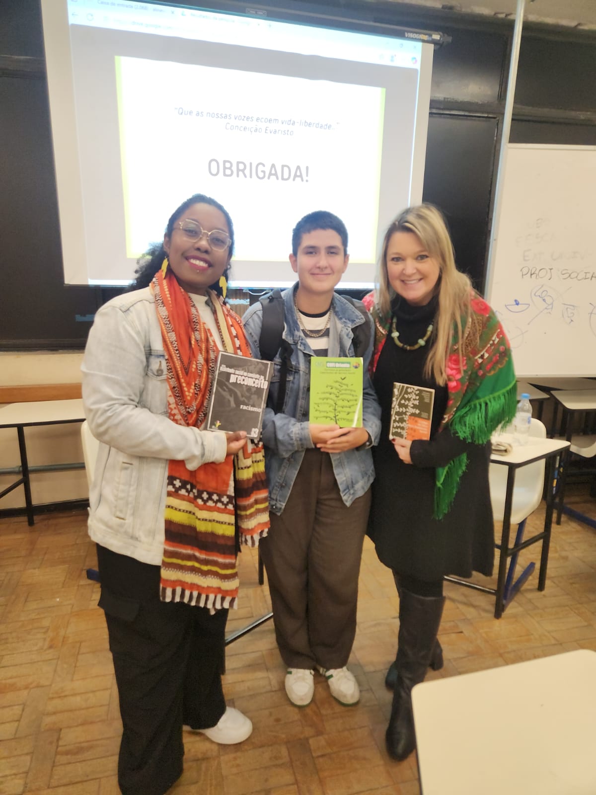 fotografia com a presença de uma mulher, negra, com cabelos crespos presos; de uma pessoa não binária, branca, de cabelos curtos, pretos; e de uma mulher branca de cabelos loiros, lisos. Elas estão dentro de uma sala de aula e seguram livros sobre o Serviço Social.