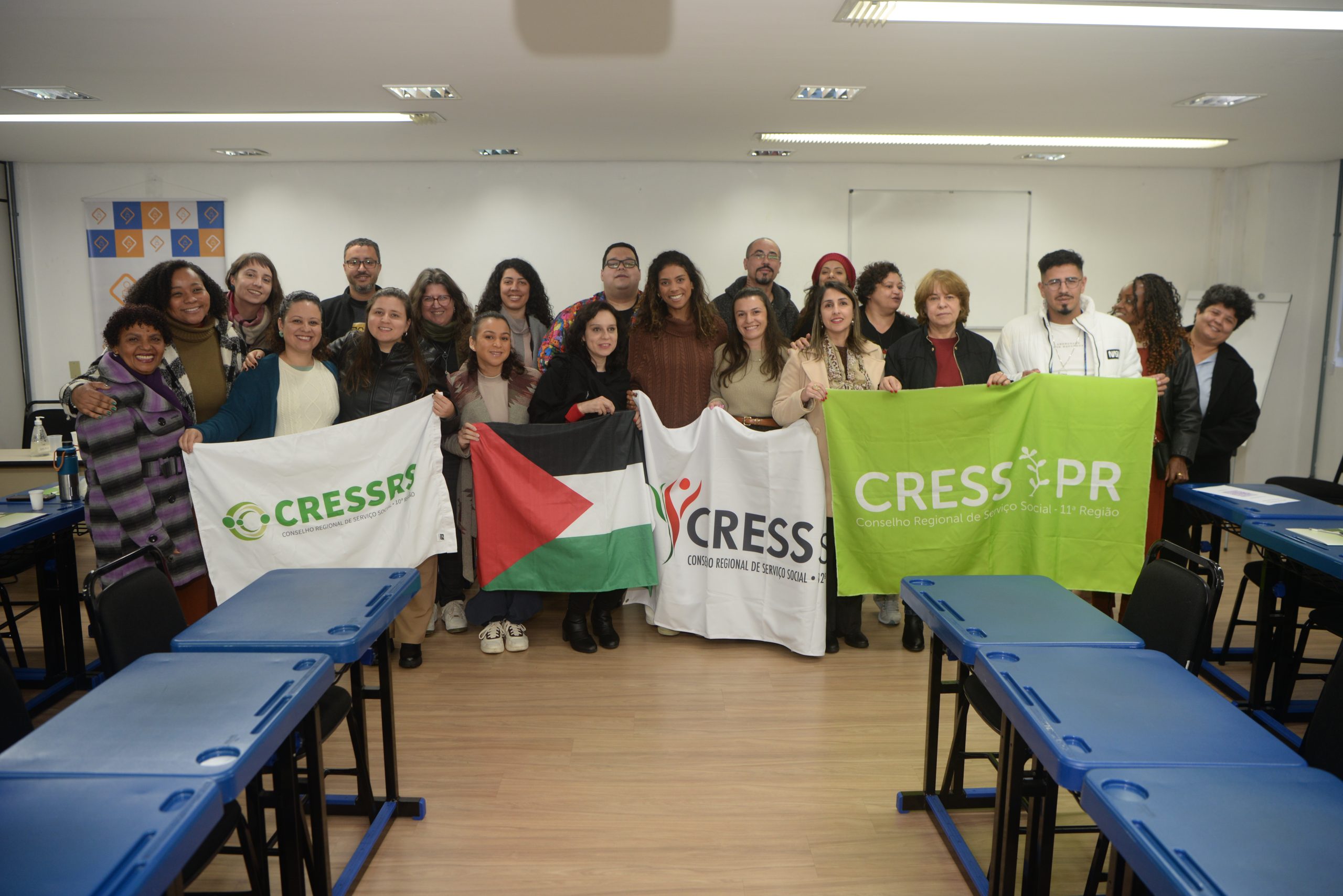 Fotografia com 21 pessoas, reunidas no salão da FAMURS, segurando uma bandeira da Palestina e bandeiras dos Cress RS, SC, PR.