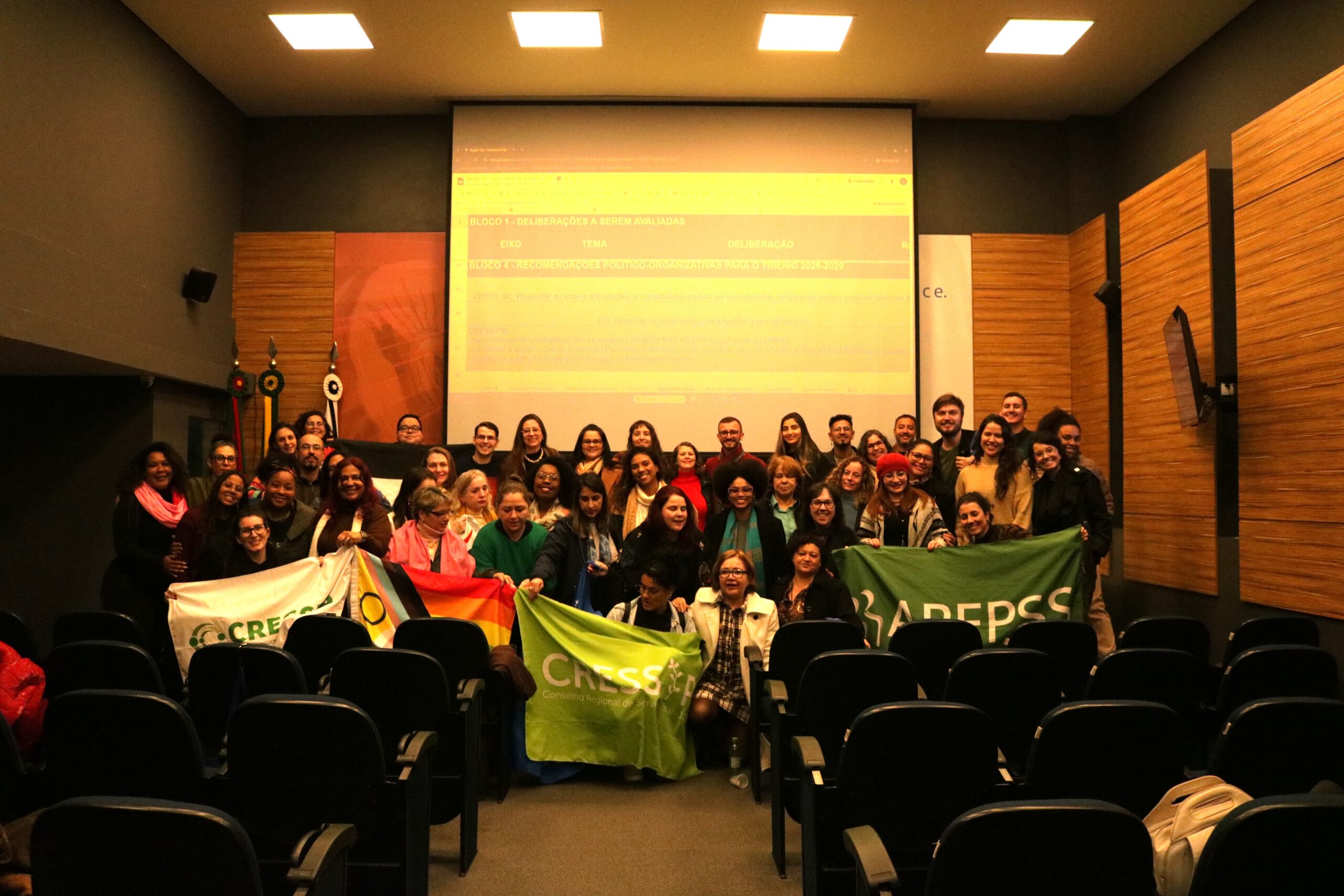 Um grupo com dezenas de pessoas posa para foto segurando bandeiras dos CRESS RS, SC, PR, ENESSO, ABEPSS, CFESS.