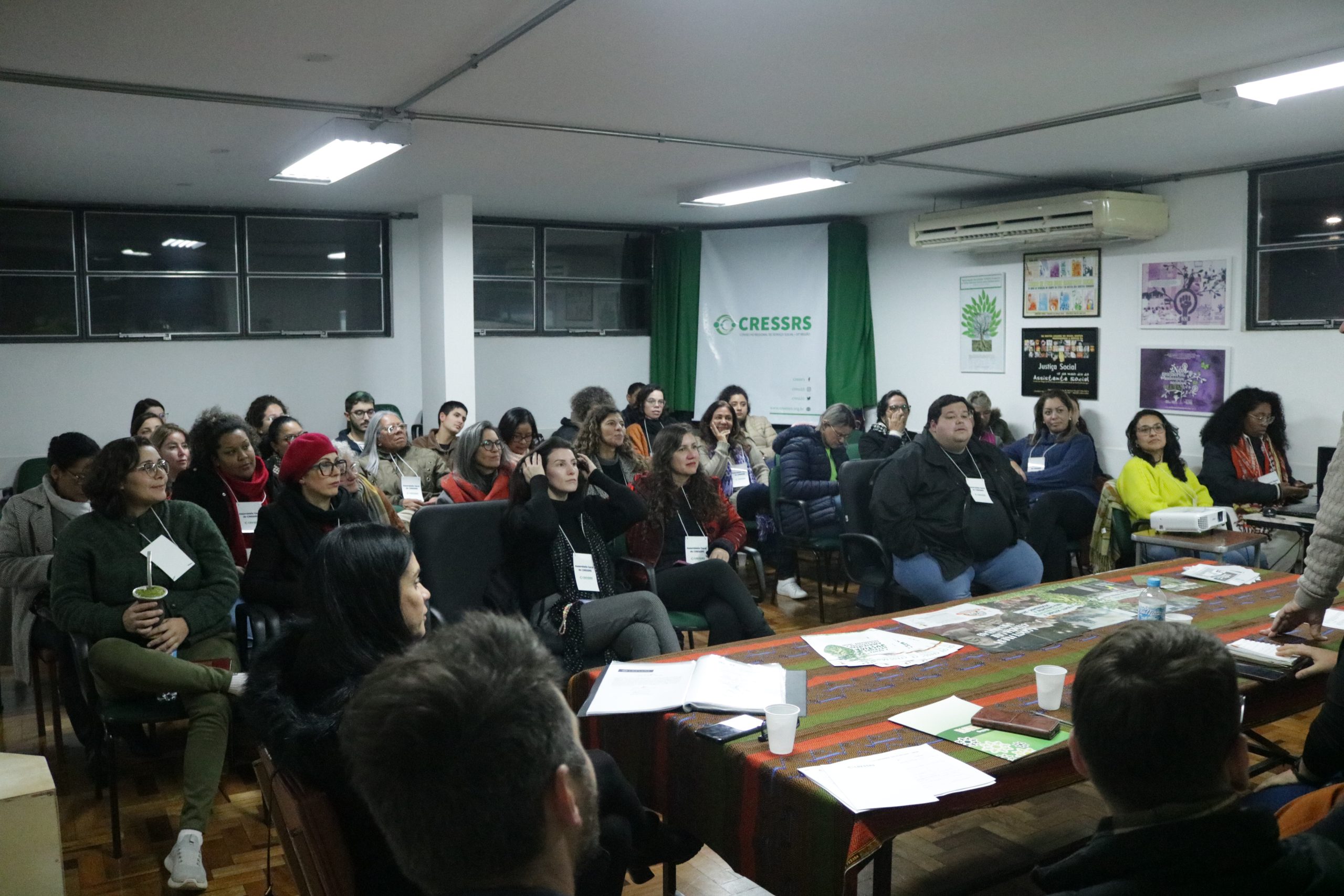 Fotografia do auditório do CRESSRS, com as/os assistentes sociais presentes para a assembleia, sentados em filas. São cerca de 30 pessoas. Ao fundo do auditório, um banner escrito CRESSRS.