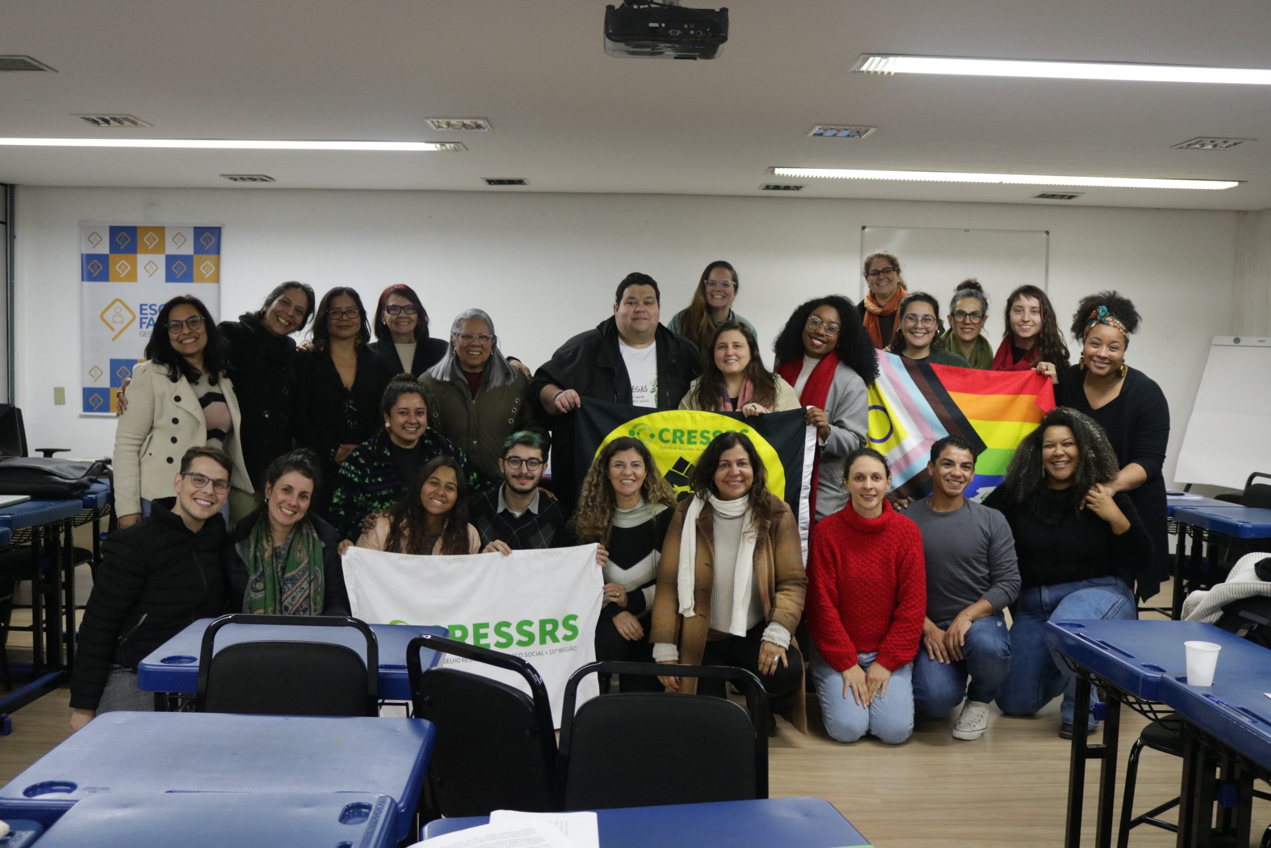 fotografia com 24 pessoas reunidas no auditório da FAMURS. Algumas pessoas seguram bandeiras do CRESSRS referentes à luta lgbtqia+ e antirracista.