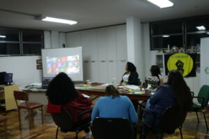 Cinco mulheres, sentadas em roda, olham para um telão onde é transmitida reunião online com mais de 15 pessoas.