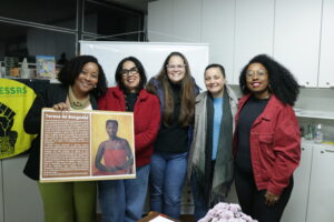 Cinco mulheres, três brancas e duas pretas, posam de pé enquanto uma delas segura um banner com a foto de Tereza de Benguela e um texto com descrição de quem foi ela. Elas estão no auditório do CRESSRS.