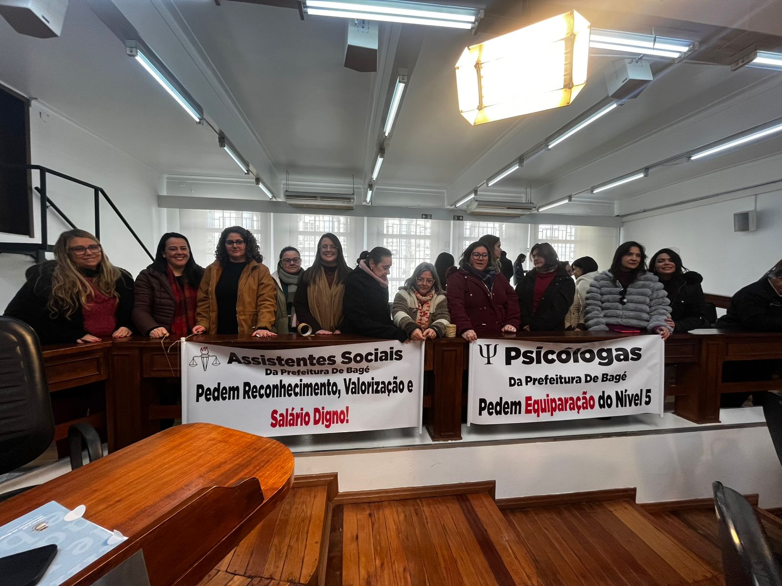 11 mulheres, brancas, com roupas e casacos de inverno, seguram faixas escrito "Assistentes Sociais da Prefeitura de Bagé pedem reconhecimento, valorização e salário digno" e "Psicólogas da Prefeitura de Bagé pedem equiparação do Nível 5".