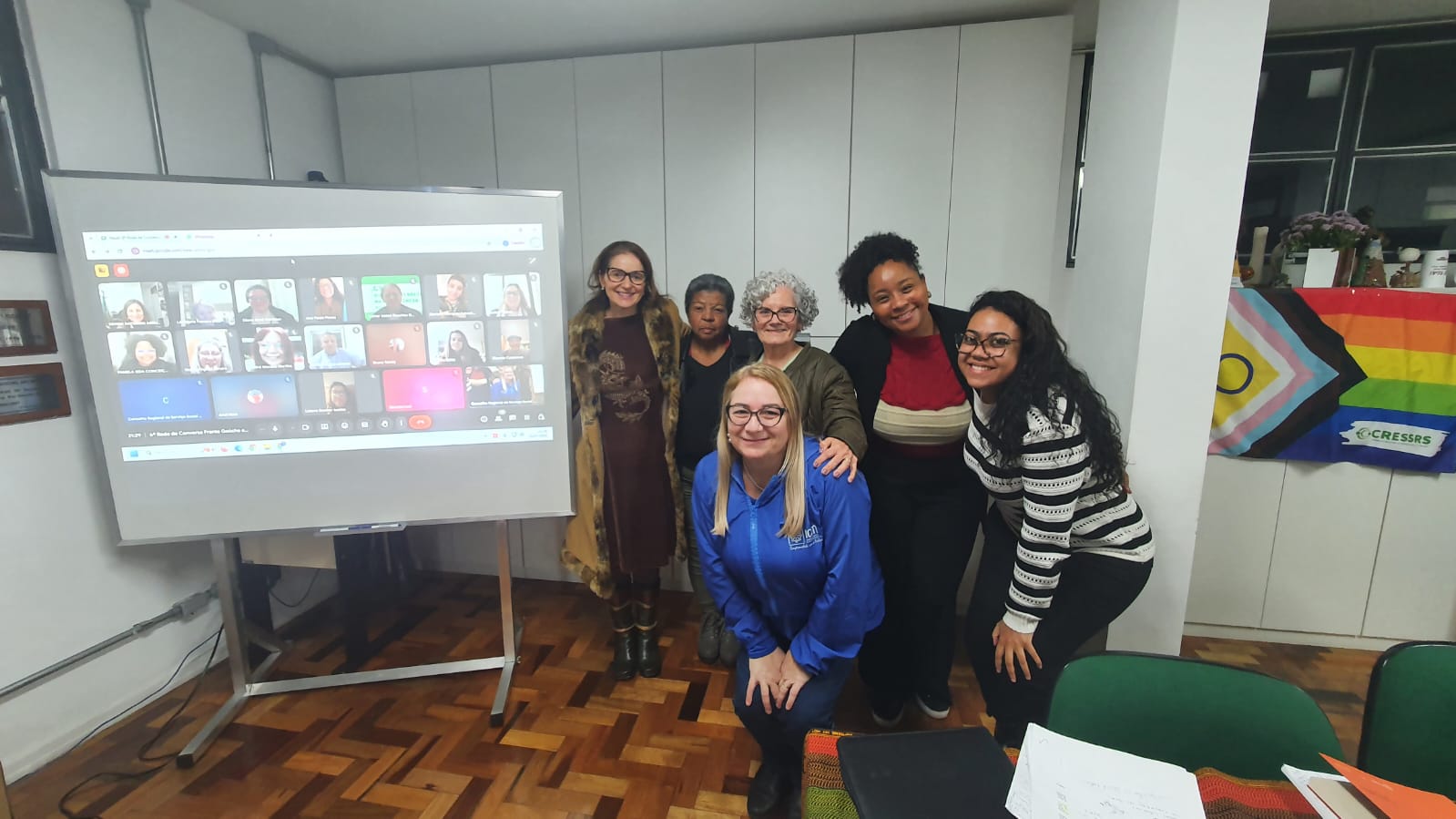 Seis mulheres posam ao lado de um telão com reunião online onde tem mais de 30 pessoas presentes.