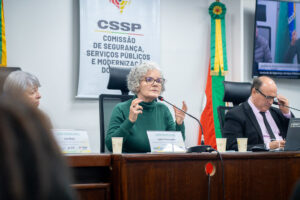 A assistente social Leila fala no microfone e gesticula, sentada em uma mesa.
