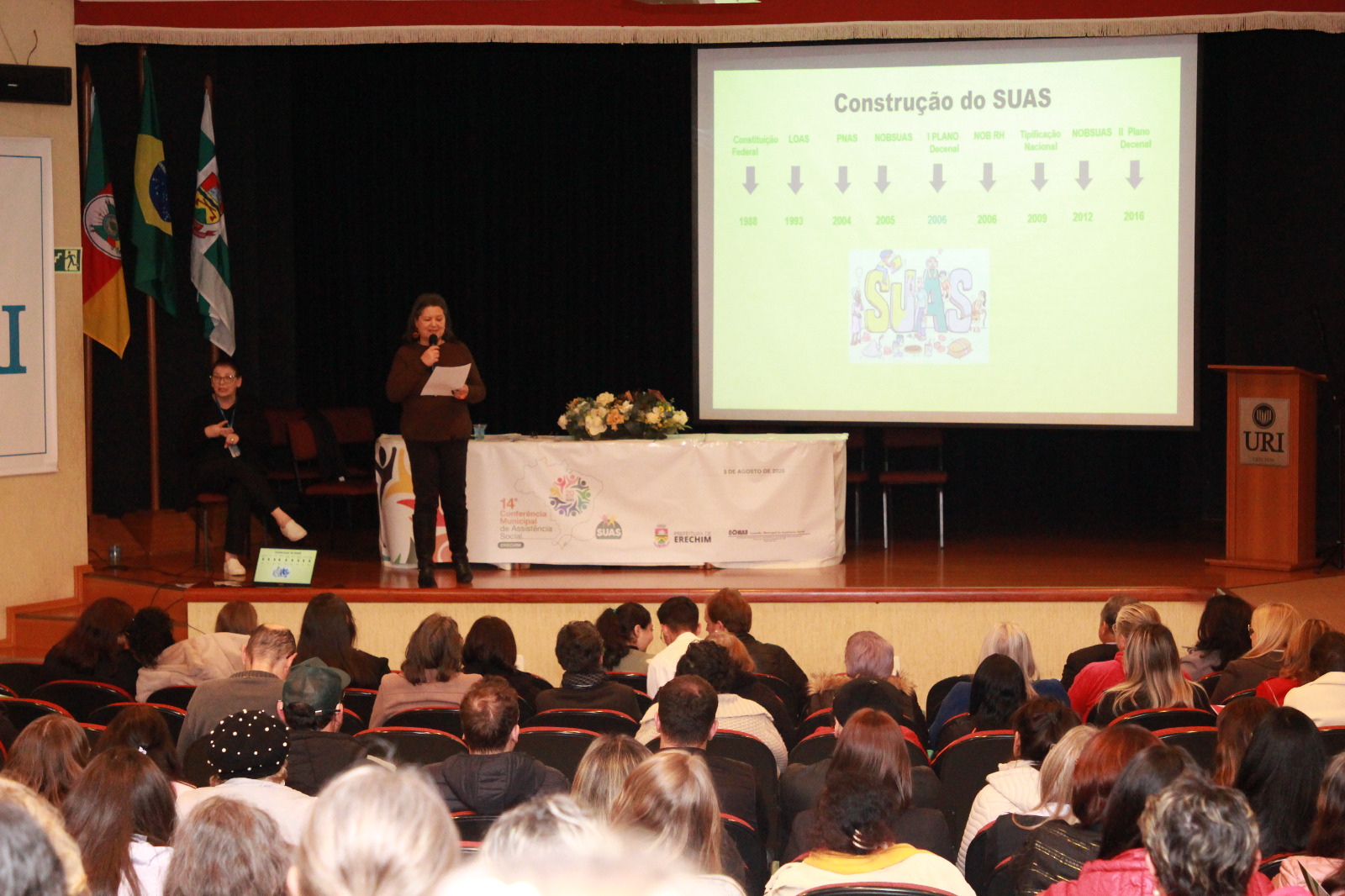 Conselheira do CRESSRS, mulher, branca, fala em cima de um palco e com microfone para plateia. Ao fundo, telão exibe apresentação.