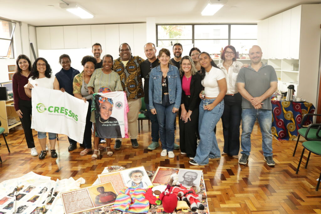 15 pessoas no auditório do CRESSRS, segurando uma bandeira do conselho. Cinco pessoal negras, 10 mulheres e cinco homens. 