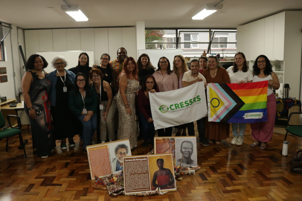 Fotografia com 17 pessoas no auditório do CRESSRS, segurando bandeiras do conselho. Três negras/os e dois homens. 