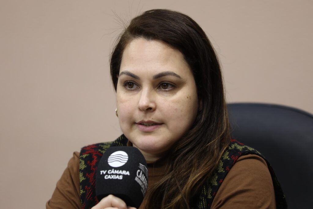 Fotografia com close de mulher, branca, cabelos castanhos e lisos falando no microfone.