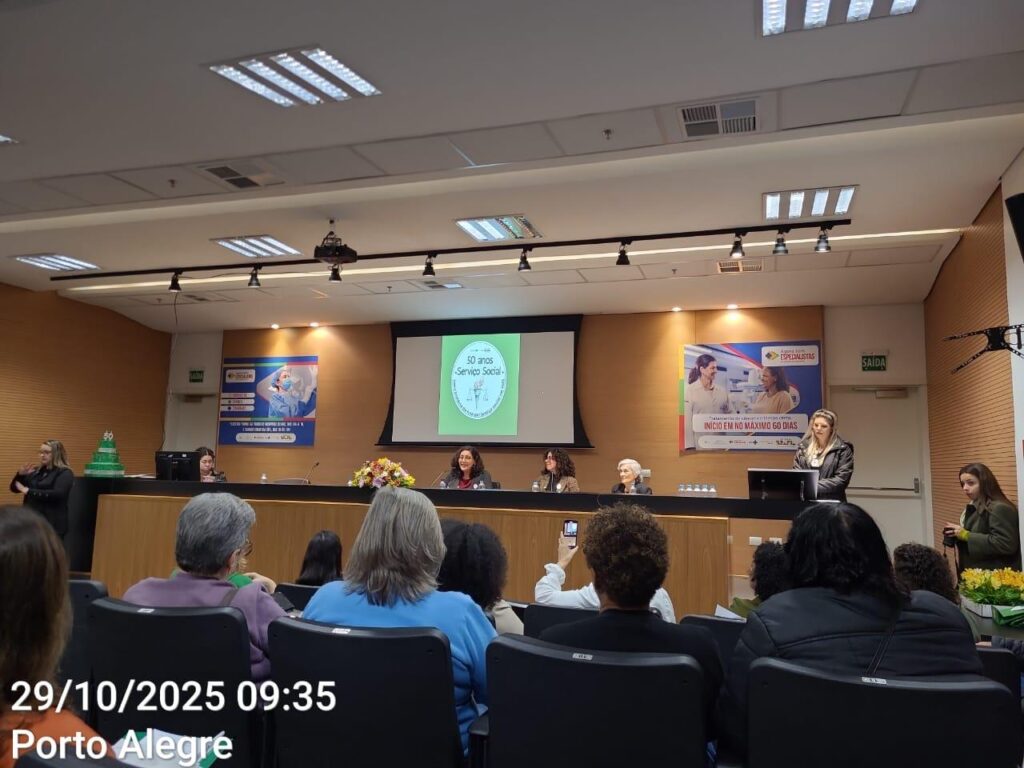 Um auditório onde três mulheres estão sentadas em uma bancada, e uma delas fala ao microfone. Atrás, é exibida uma apresentação no telão. 