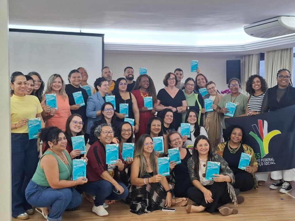 31 pessoas, maioria mulheres, reunidas segurando um livro do curso Ética em Movimento, ao lado uma bandeira do CFESS.
