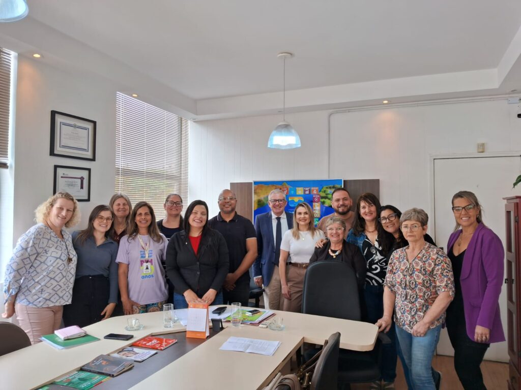 12 mulheres, brancas, de meia idade, um homem negro, dois homens brancos, posam para foto em uma sala com mesa e paredes brancas. 
