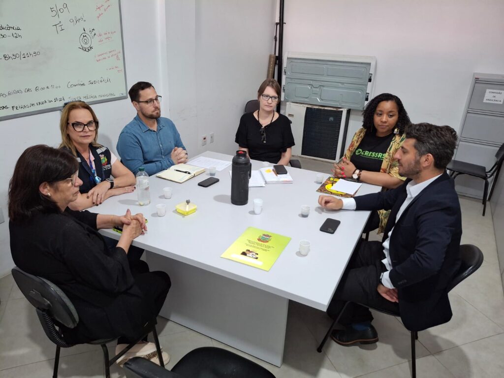Pessoas sentadas em roda, ao redor de uma mesa. Cinco mulheres, uma delasé negra. Dois homens brancos.