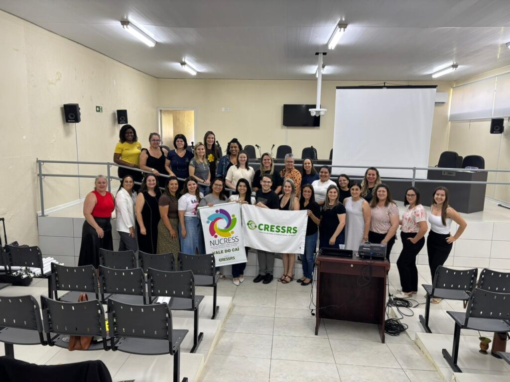 Fotografia com 27 mulheres e 01 homem, em auditório, com telão ao fundo. Elas seguram um banner no NUCRESS Vale do Caí e uma bandeira verde e branca do CRESSRS.