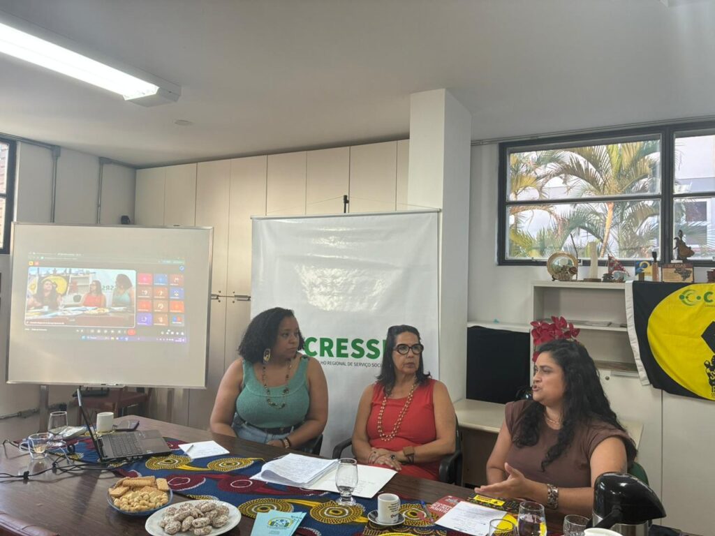 Uma mulher negra e duas mulheres indígenas reunidas no auditório do CRESSRS, com telão ao fundo com reunião no Meet e banner do CRESSRS.