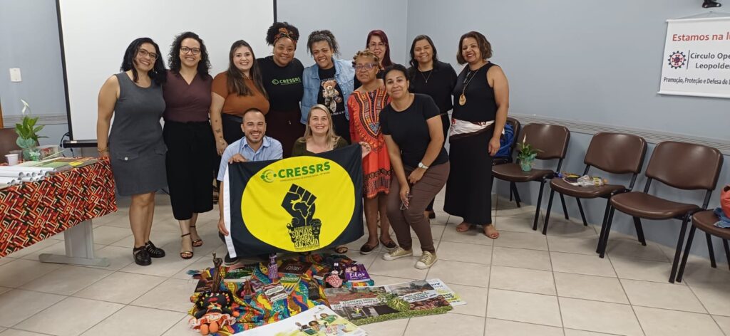 11 mulheres brancas e negras e 01 homem reunidos segurando a bandeira do CRESSRS de combate ao racismo.