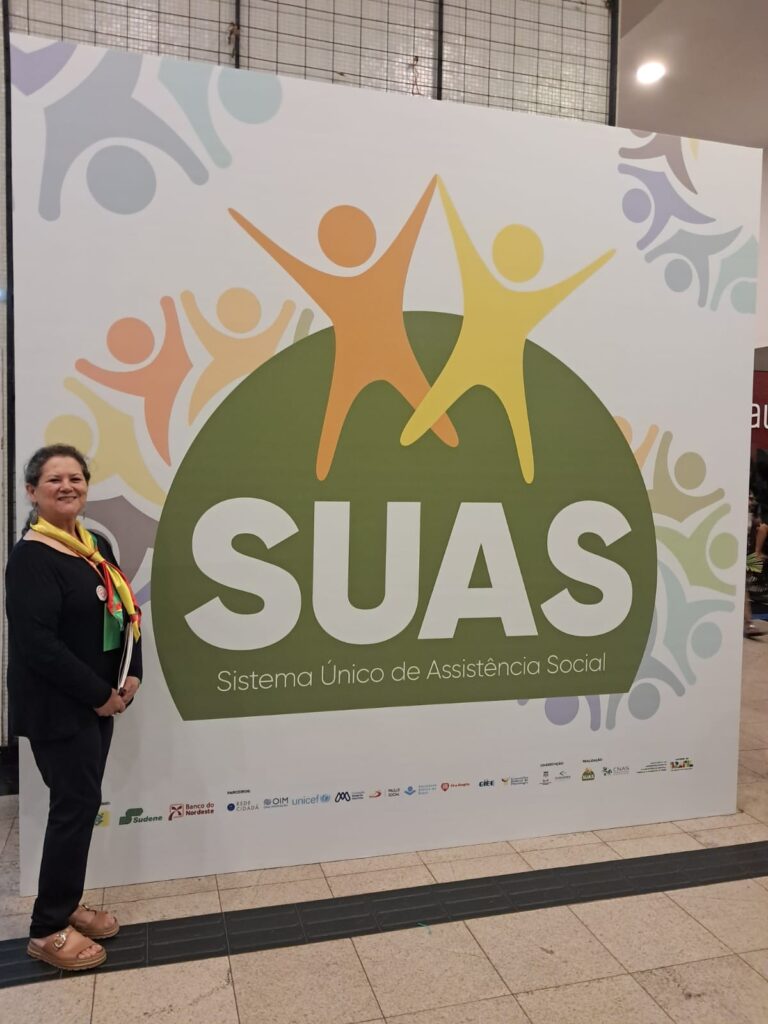 Mulher, branca, posa em frente a um painel escrito SUAS Sistema Único de Assistência Social.
