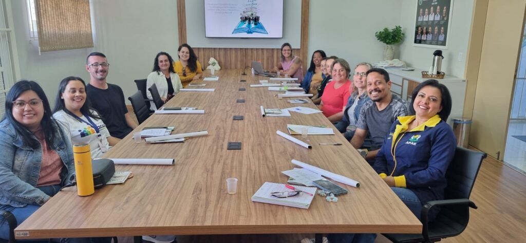 Foto com 11 mulheres e dois homens, sentados ao redor de uma grande mesa retangular, com cartazes e blocos em suas frentes. 