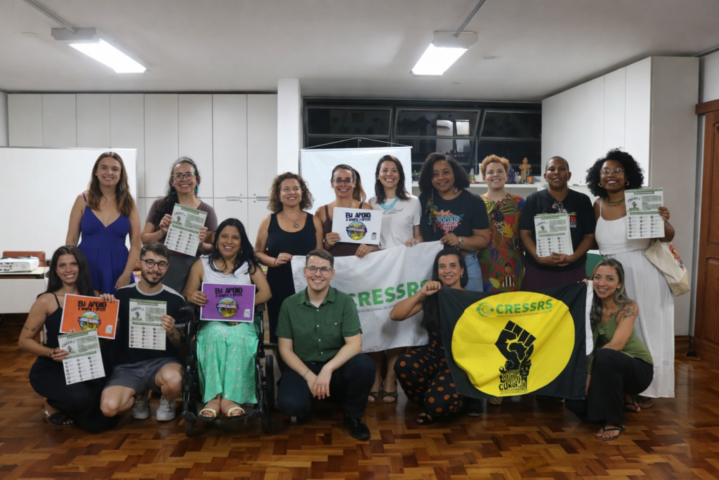 Registro fotográfico do evento, com 17 pessoas reunidas posando para a foto. Elas seguram cartazes das chapas que concorrem ao CRESS-RS e e ao CFESS e seguram uma bandeira de combate ao racismo, preta e amarela, com um punho cerrado no centro e com a logo do CRESS-RS. A foto foi tirada no auditório do CRESS-RS no final do debate.