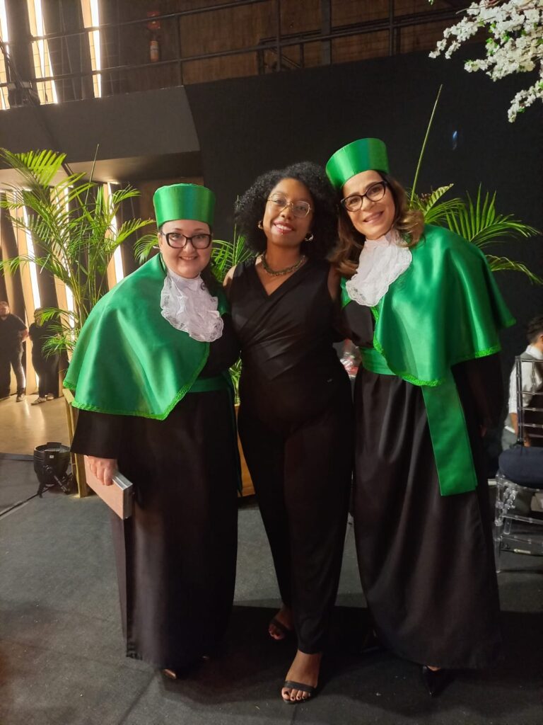 Fotografia da conselheira do CRESSRS, mulher, negra, vestida com macacão preto, entre duas formandas, mulheres, brancas, com toga verde.