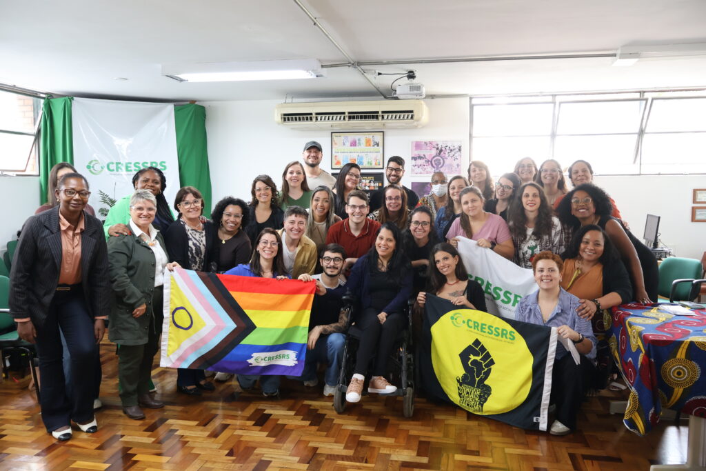 Fotografia com 32 pessoas, maioria mulheres brancas e cinco negras, 4 homens, 1 mulher com deficiência física. Eles estão no auditório do CRESSRS e seguram bandeiras do CRESSRS em alusão a luta antirracista e LGBTQIA+.