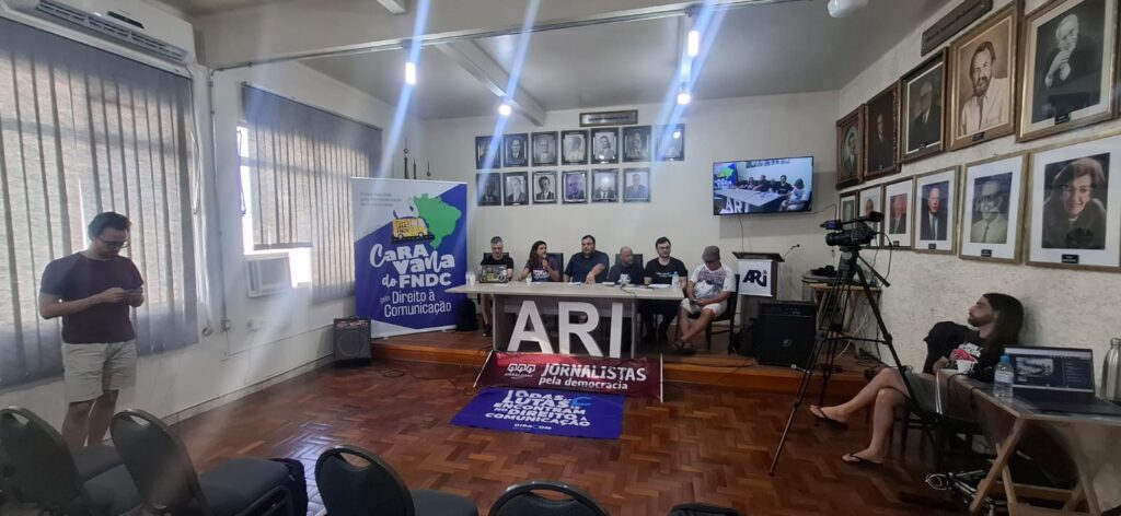 Foto no auditório da ARI, com banner da caravana do FNDC ao lado de uma mesa, onde estão sentados cinco homens e uma mulher.