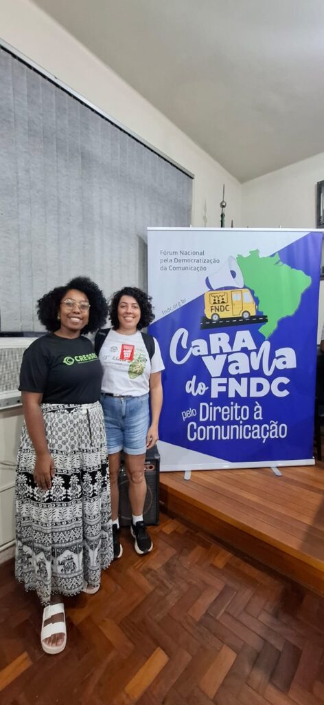 Conselheira do CRESSRS, mulher, negra e assessora de comunicação do CRESSRS, mulher parda, ao lado do banner da caravana do FNDC.