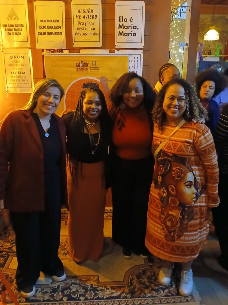 Três mulheres pretas e uma branca, reunidas em um bar, com parede cheia de cartazes ao fundo.