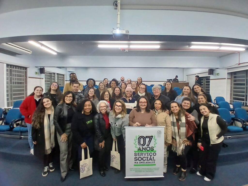 Fotografia com 35 pessoas, maioria mulheres, negras e brancas, reunidas na UniLaSalle segurando um banner escrito 07 anos Serviço Social na Unilasalle!