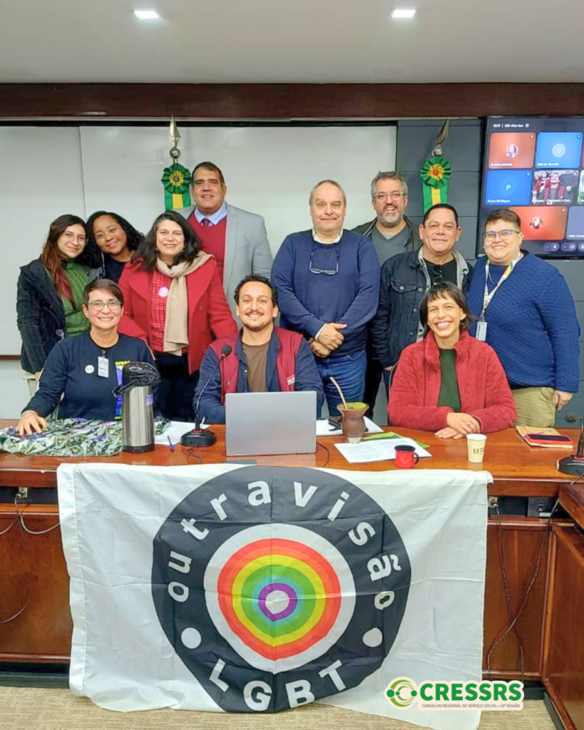 Fotografia com cinco mulheres brancas, uma mulher negra e cinco homens brancos, atrás de uma mesa. Na frente da mesa, está uma bandeira escrito Outra Visão LGBT. 