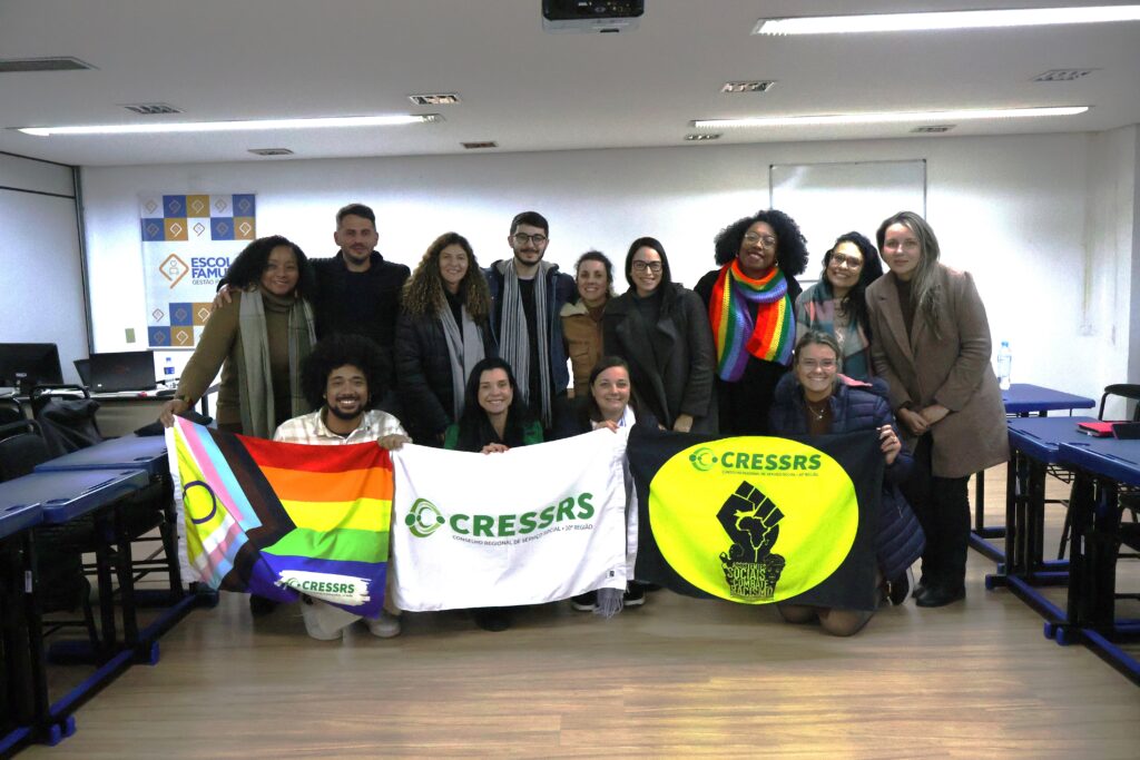 Fotografia com 13 pessoas, mulheres e homens, três pessoas negras, reunidas no auditório da FAMURS. Elas seguram três bandeiras do CRESSRS: uma branca com a logo verde, uma LGBTQIAPN+ colorida com as cores do arco-íris e uma antirracista preto e amarela.