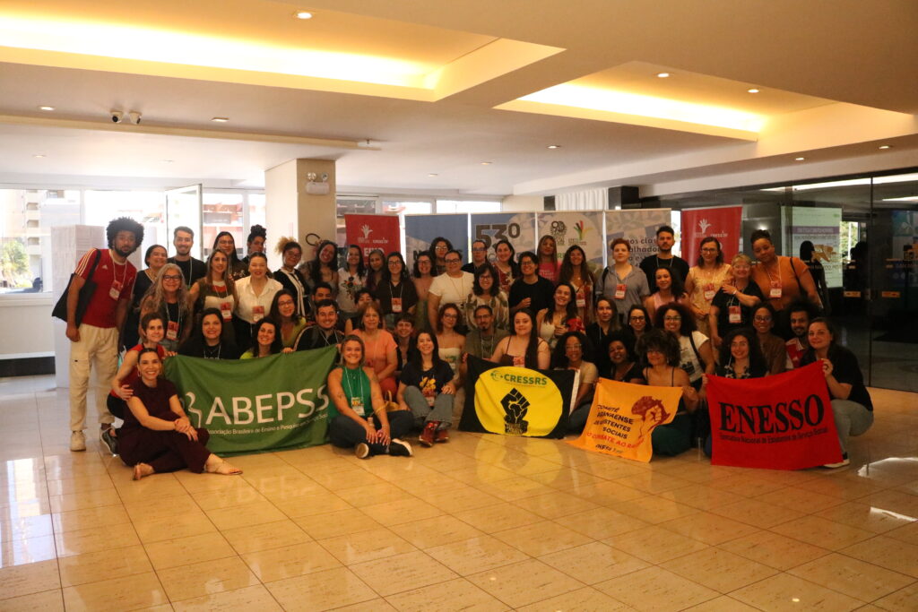 Fotografia com 53 pessoas reunidas em frente aos banners do evento, segurando bandeiras da ABEPSS, CRESSRS, ENESSO.