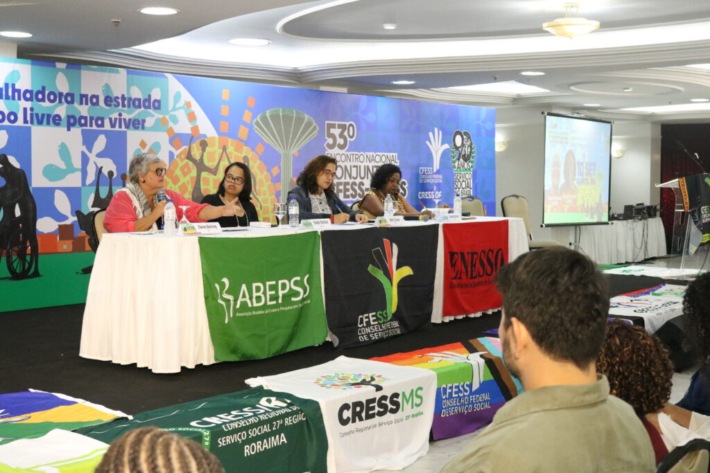 Fotografia de mesa de debates com uma mulher branca e três mulheres negras. Em frente à mesa bandeiras da ABEPSS, CFESS e ENESSO.