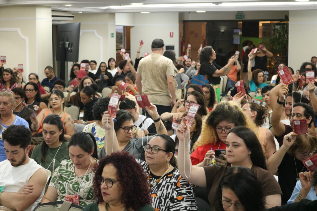 Auditório com muitas mulheres e alguns homens levando crachás para o alto em momento de votação.