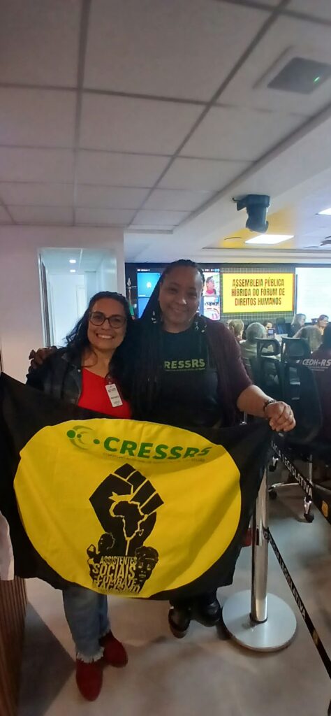 Foto vertical de mulher parda e mulher negra segurando uma bandeira do CRESSRS em alusão a luta antirracista. Elas esttão em auditório na Assembleia Legislativa.