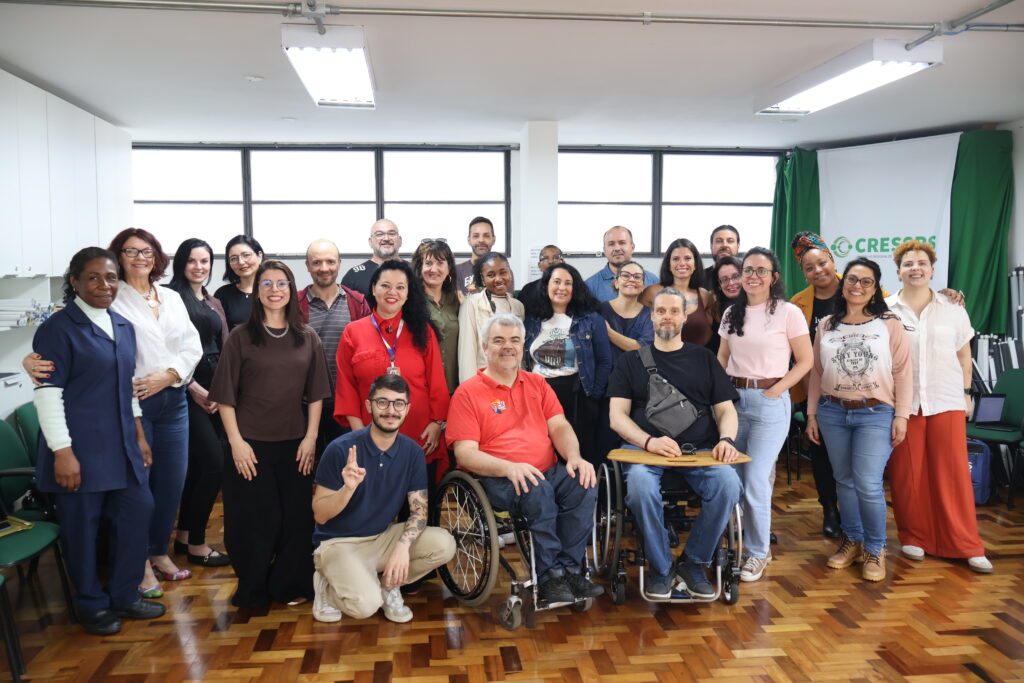 Fotografia com 11 mulheres brancas, 5 mulheres negras e 8 homens brancos, dois deles em cadeiras de rodas. Eles estão de pé e sentados em cadeiras de rodas, no auditório do CRESSRS.