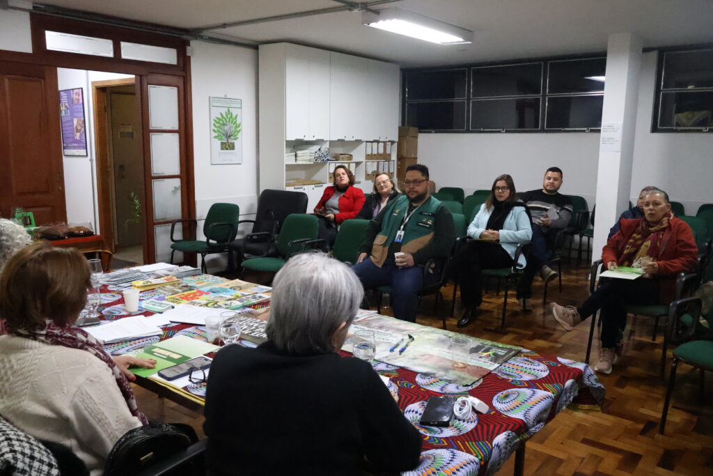 Foto do auditório do CRESSRS com dois homens de cabelos castanhos e cinco mulheres brancas sentadas na plateia enquanto uma mulher de cabelos grisalhos está de frente para estas pessoas, sentada em uma cadeira com uma mesa em sua frente.